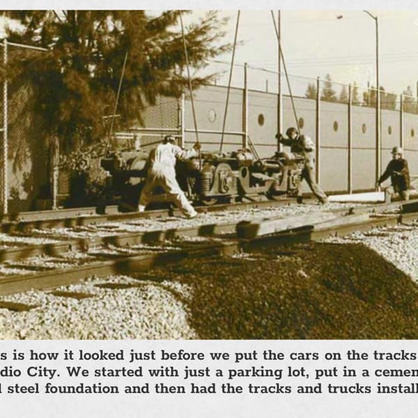 Three workers installing train trucks onto tracks in Studio City, with a fence and building in the background.