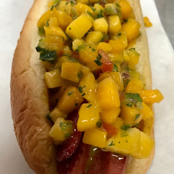 Hot dog in a bun topped with vibrant yellow mango salsa, green herbs, and red peppers.