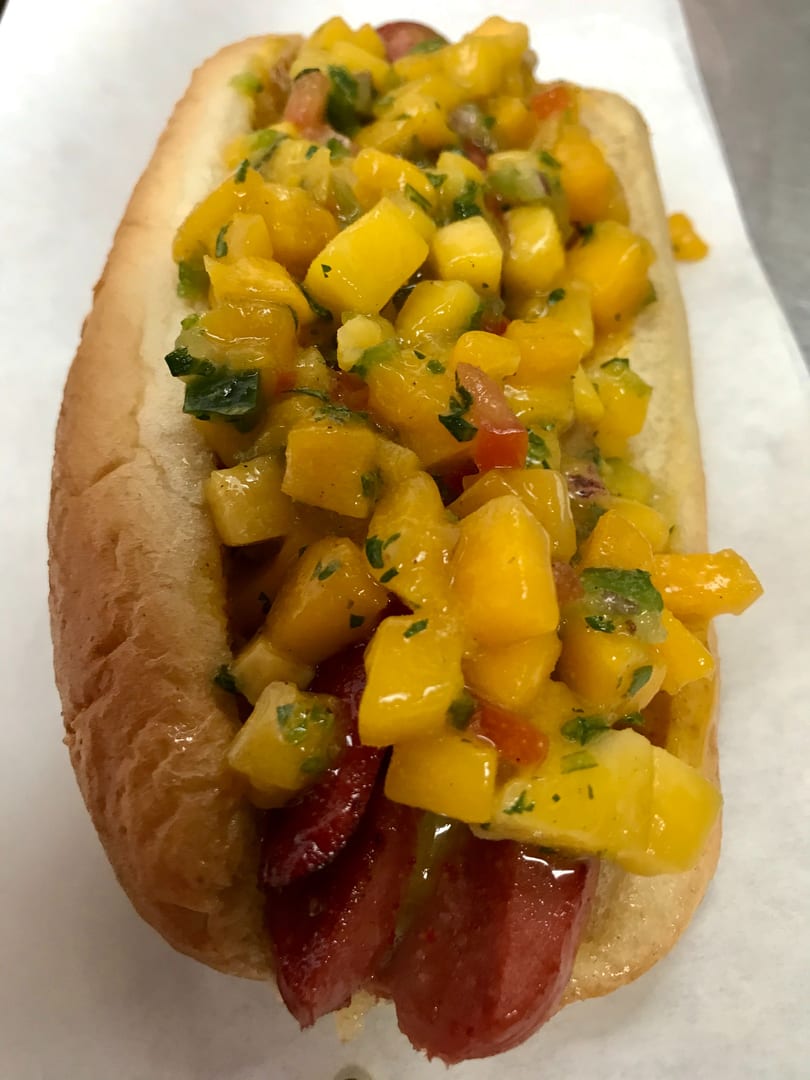 Hot dog in a bun topped with vibrant yellow mango salsa, green herbs, and red peppers.