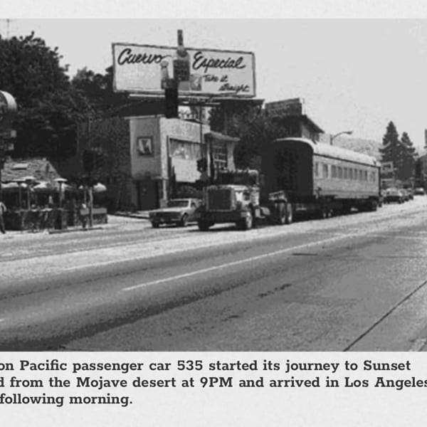 Black and white photo of a train car on a flatbed truck driving down a street.