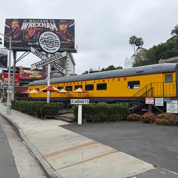 Carney's restaurant, a yellow train car with red umbrellas, sits beside a street with billboards.