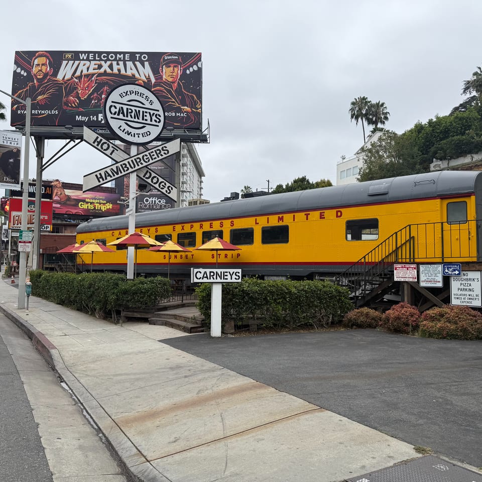 Carney's restaurant, a yellow train car with red umbrellas, sits beside a street with billboards.