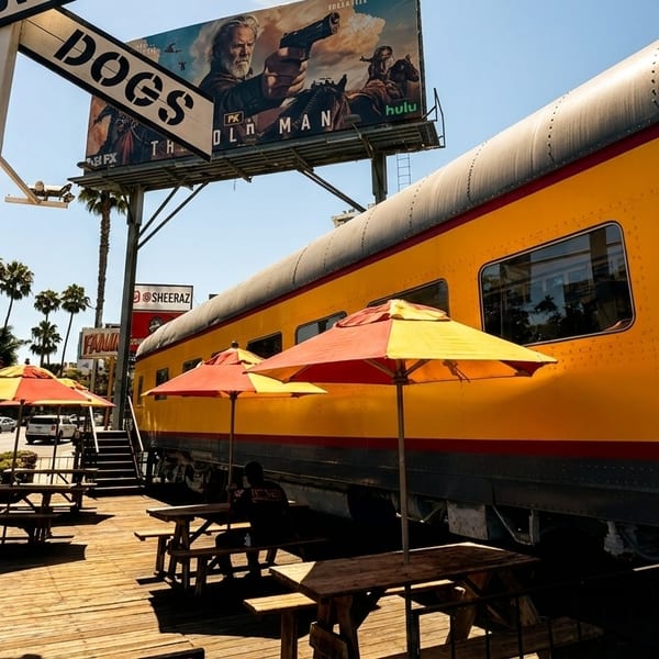 Outdoor dining area with picnic tables and umbrellas next to a yellow train car restaurant.