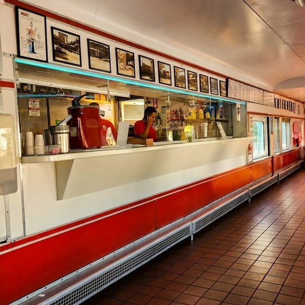 A long, narrow diner interior with a counter, red booths, and a person seated at a table.