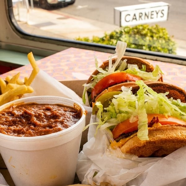 Carney's chili, fries, and burgers with lettuce and tomato, on a patterned table by a window.